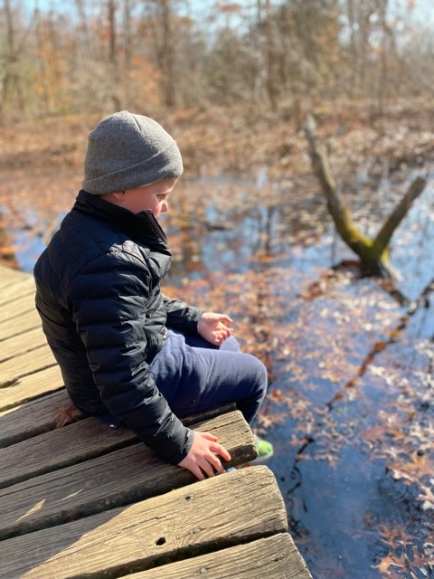 A young boy sits on the boardwalk looking at a pond